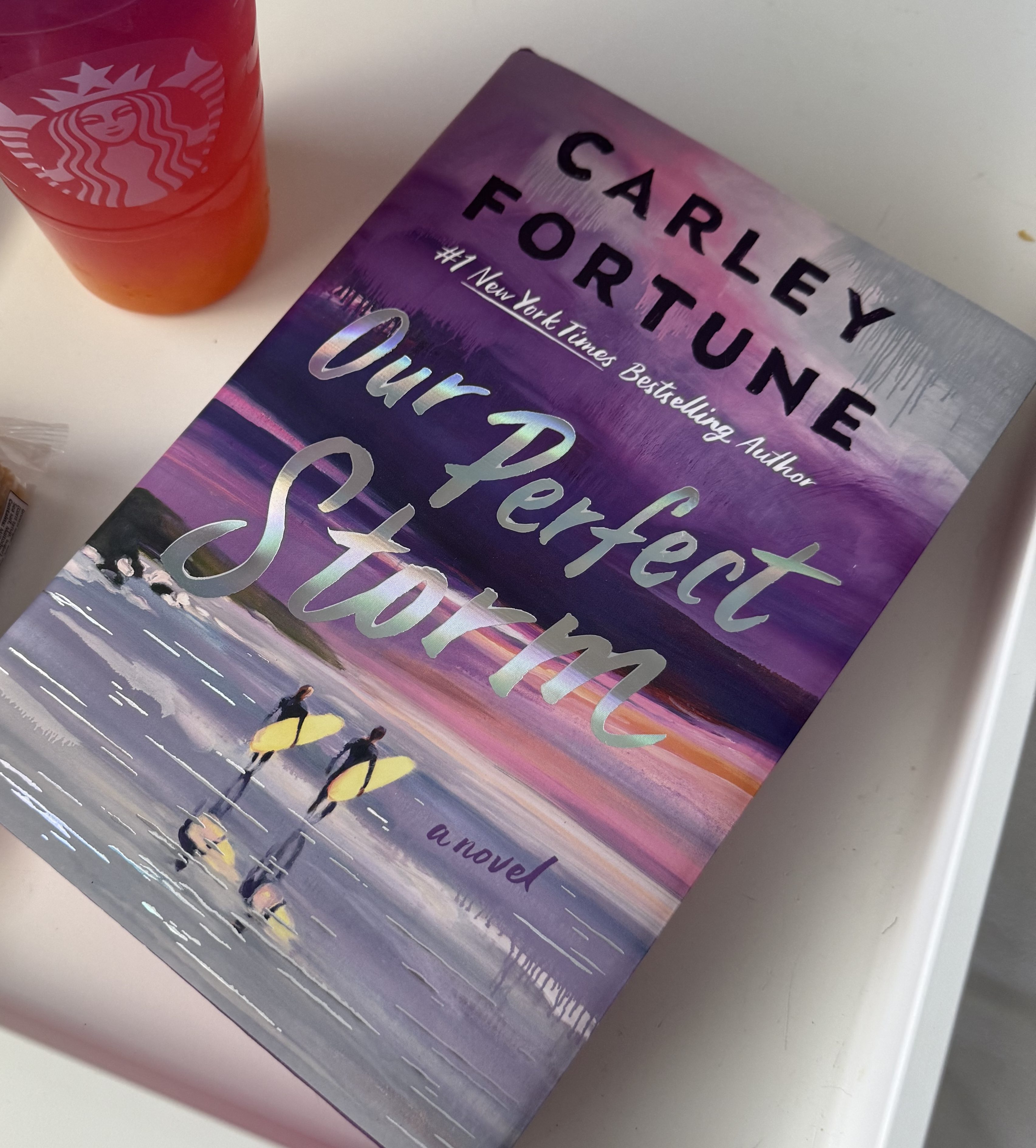 A close-up of the book 'Our Perfect Storm' by Carley Fortune, featuring a colorful cover with surfers and a sunset, alongside a Starbucks drink and a snack.