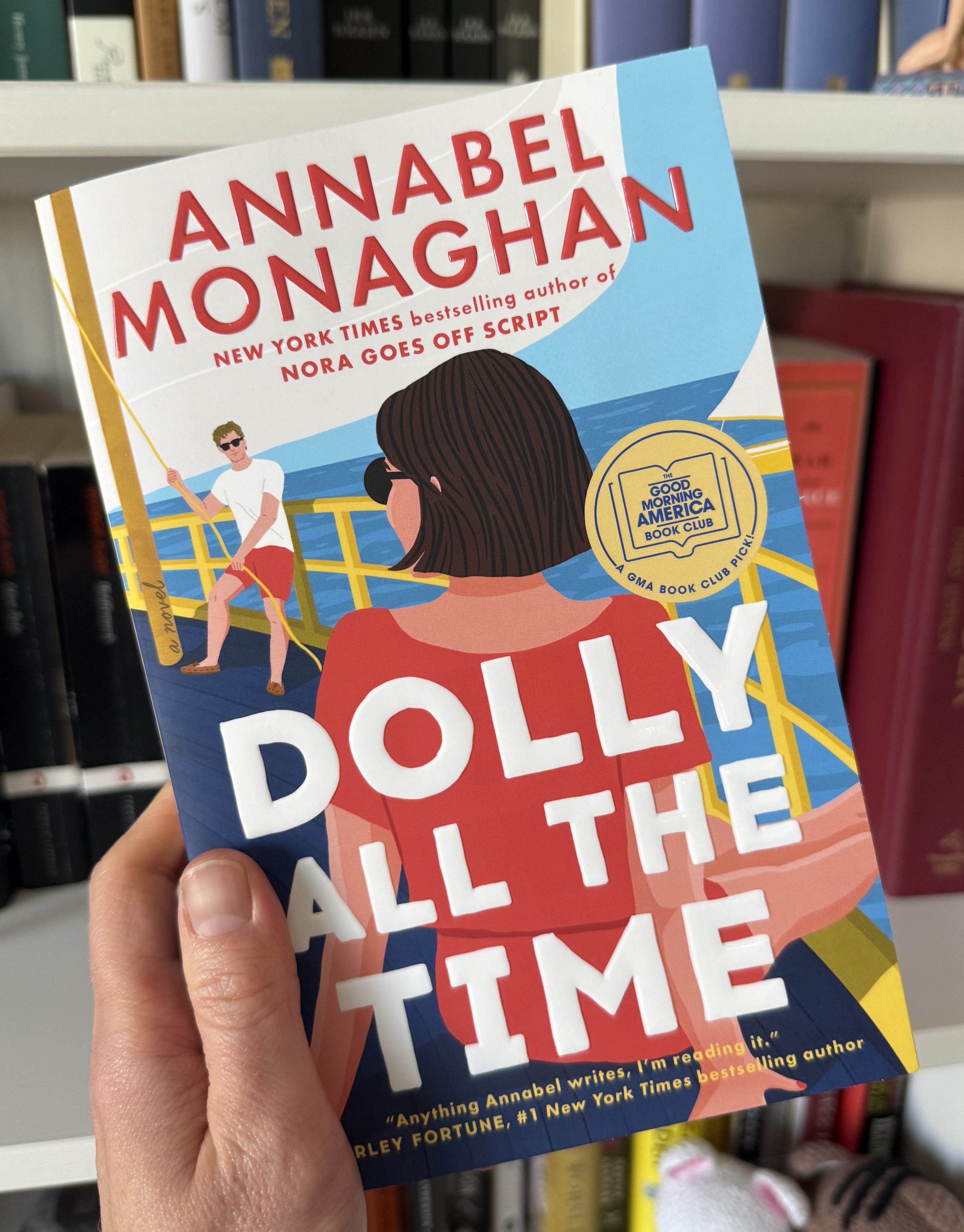 A person holding a book titled 'Dolly All the Time' by Annabel Monaghan, with a colorful cover illustration featuring two characters on a boat.
