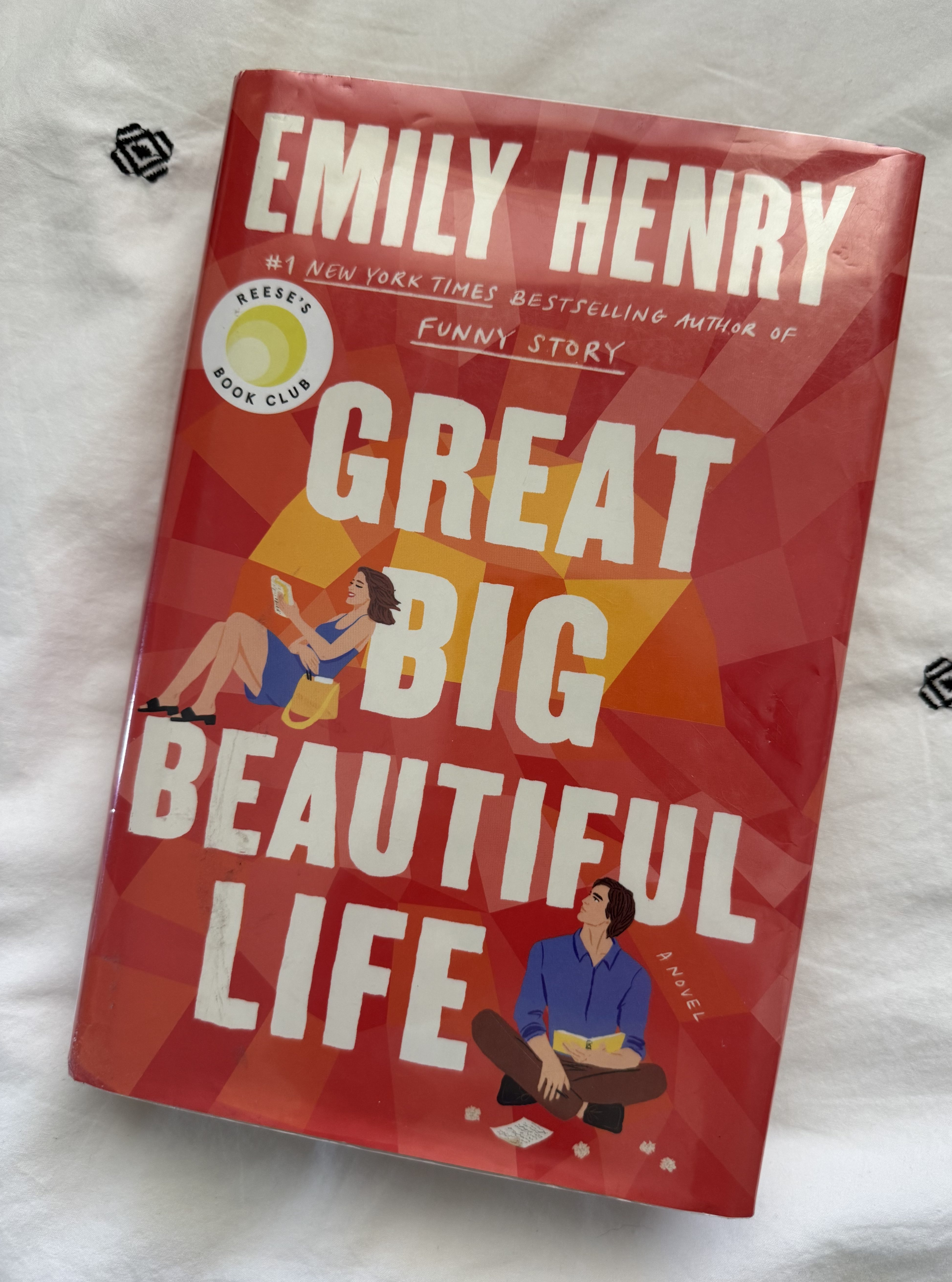 A close-up view of the book cover for 'Great Big Beautiful Life' by Emily Henry, featuring a vibrant red background with illustrations of a woman and a man reading. The book is also noted as a selection for Reese's Book Club.